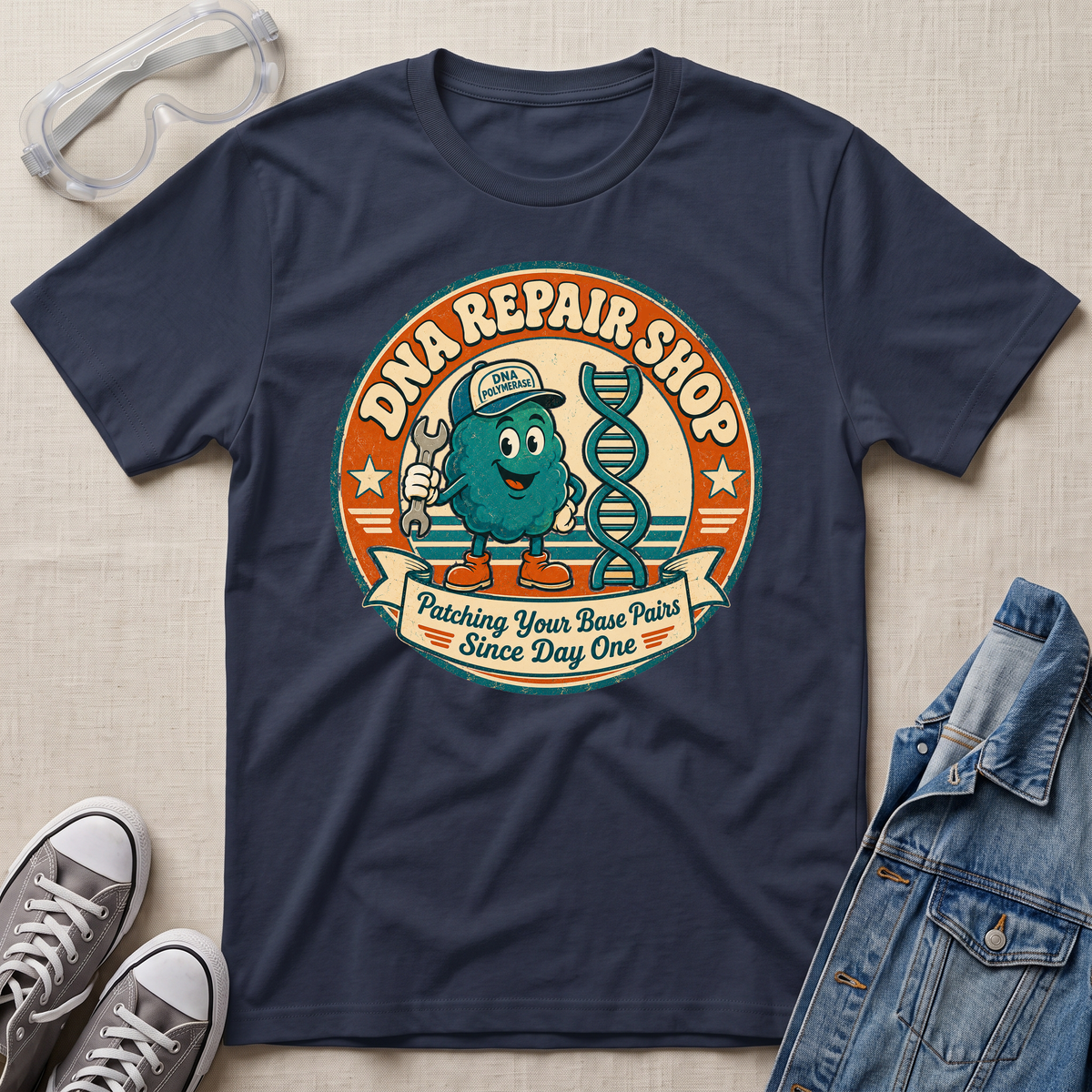 A navy blue graphic t-shirt laid flat on a beige fabric background, styled with clear safety goggles, gray sneakers, and a denim jacket. The t-shirt features a retro circular graphic for 'DNA REPAIR SHOP' with a cartoon teal DNA helix character and the text 'Patching Your Base Pairs Since Day One'.