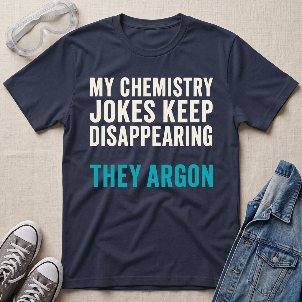 A navy blue graphic t-shirt with the text 'MY CHEMISTRY JOKES KEEP DISAPPEARING THEY ARGON' in white and teal lettering, styled in a flat lay with safety goggles, gray sneakers, and a denim jacket on a beige fabric background.