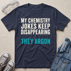 A navy blue graphic t-shirt with the text 'MY CHEMISTRY JOKES KEEP DISAPPEARING THEY ARGON' in white and teal lettering, styled in a flat lay with safety goggles, gray sneakers, and a denim jacket on a beige fabric background.