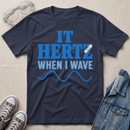 A navy blue graphic t-shirt laid flat on a textured beige fabric background, styled with safety goggles, gray sneakers, and a denim jacket. The t-shirt features the text 'IT HERTZ WHEN I WAVE' in blue and white, with a small band-aid graphic over the letter Z.