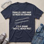 A navy blue graphic t-shirt laid flat on a beige fabric background, surrounded by styling props including safety goggles, gray sneakers, and a denim jacket. The t-shirt features a white graphic of two parallel sad lines with the text 'PARALLEL LINES HAVE SO MUCH IN COMMON' above and 'IT'S A SHAME THEY'LL NEVER MEET' below.