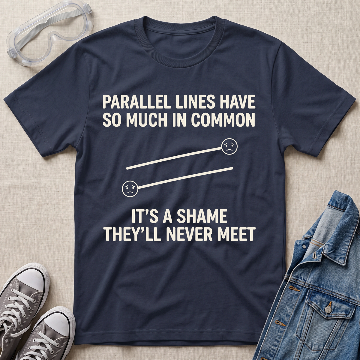 A navy blue graphic t-shirt laid flat on a beige fabric background, surrounded by styling props including safety goggles, gray sneakers, and a denim jacket. The t-shirt features a white graphic of two parallel sad lines with the text 'PARALLEL LINES HAVE SO MUCH IN COMMON' above and 'IT'S A SHAME THEY'LL NEVER MEET' below.