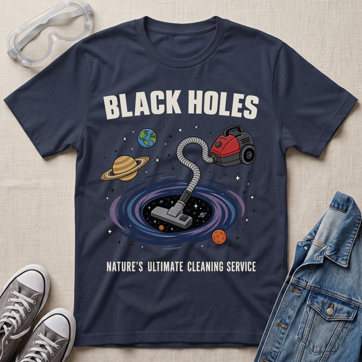 A navy graphic t-shirt laid flat on a textured fabric background, styled with safety goggles, sneakers, and a denim jacket. The t-shirt features a cartoon illustration of a vacuum cleaner falling into a black hole surrounded by planets, with the text 'BLACK HOLES' and 'NATURE'S ULTIMATE CLEANING SERVICE'.