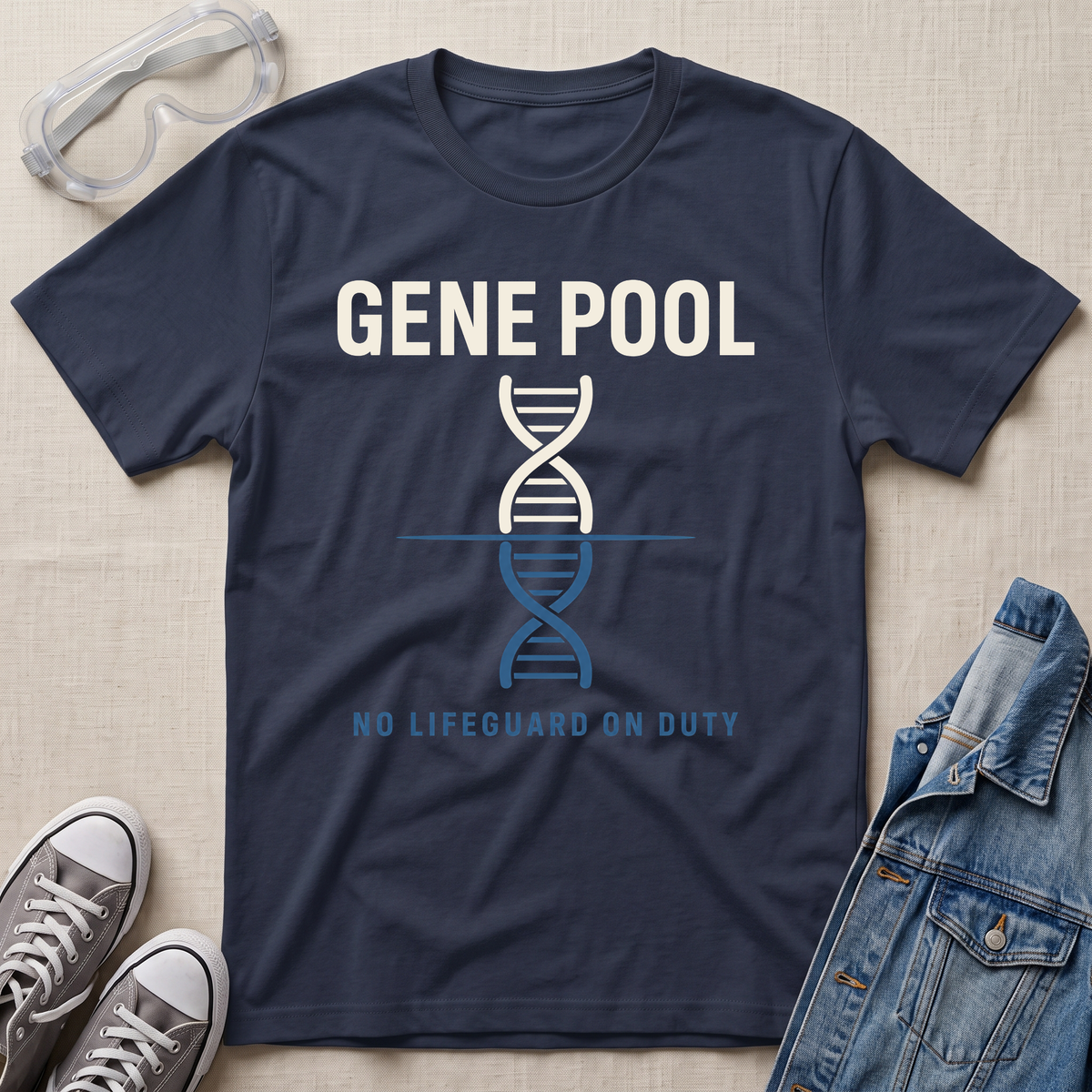 A navy blue graphic t-shirt laid flat on a light beige textured background, surrounded by lifestyle props including safety goggles, gray sneakers, and a denim jacket. The t-shirt features a white and blue DNA double helix graphic with the text 'GENE POOL' and 'NO LIFEGUARD ON DUTY'.