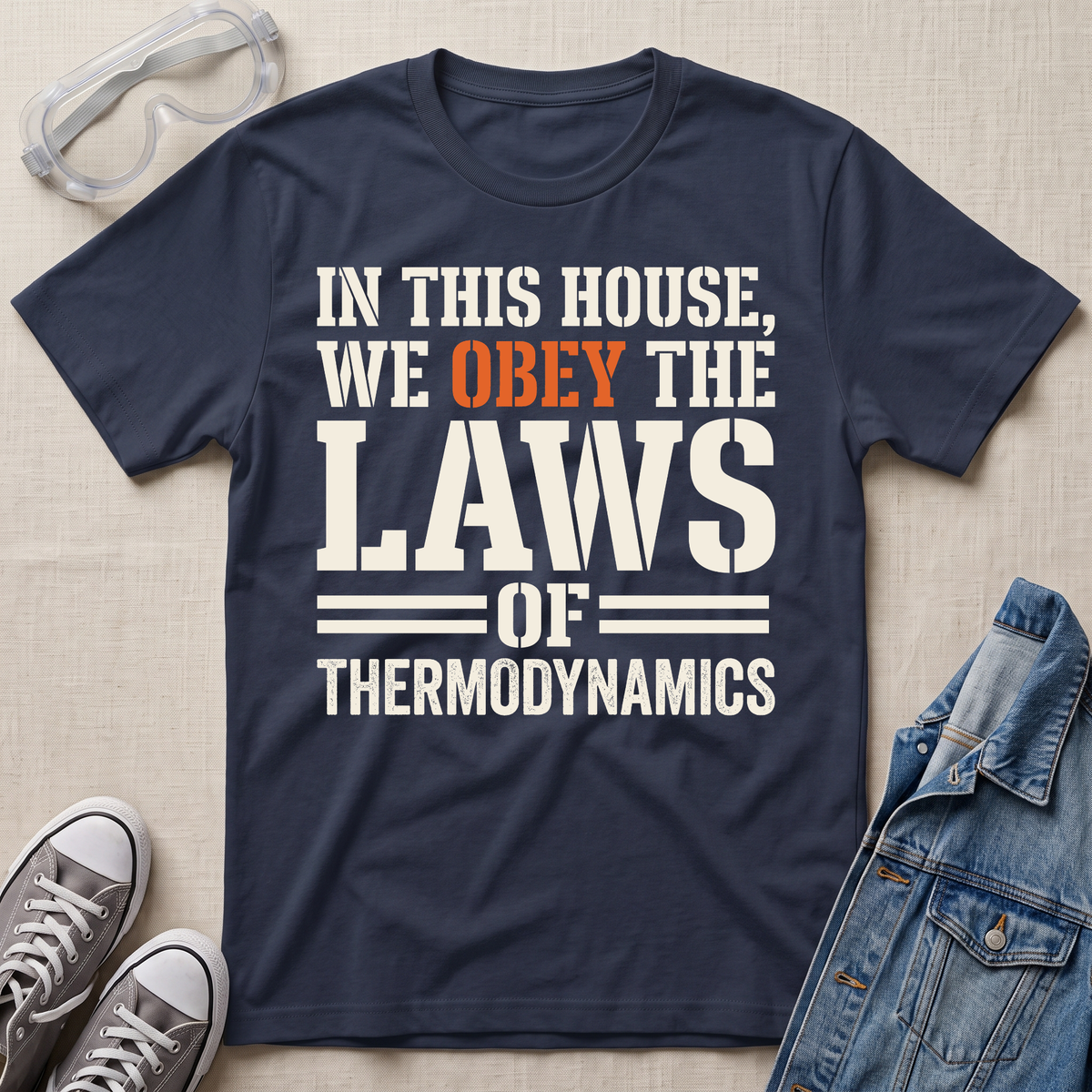 A navy blue graphic t-shirt laid flat on a beige fabric background, styled with safety goggles, gray sneakers, and a denim jacket. The t-shirt features the text 'IN THIS HOUSE, WE OBELY THE LAWS OF THERMODYNAMICS' in white and orange lettering.