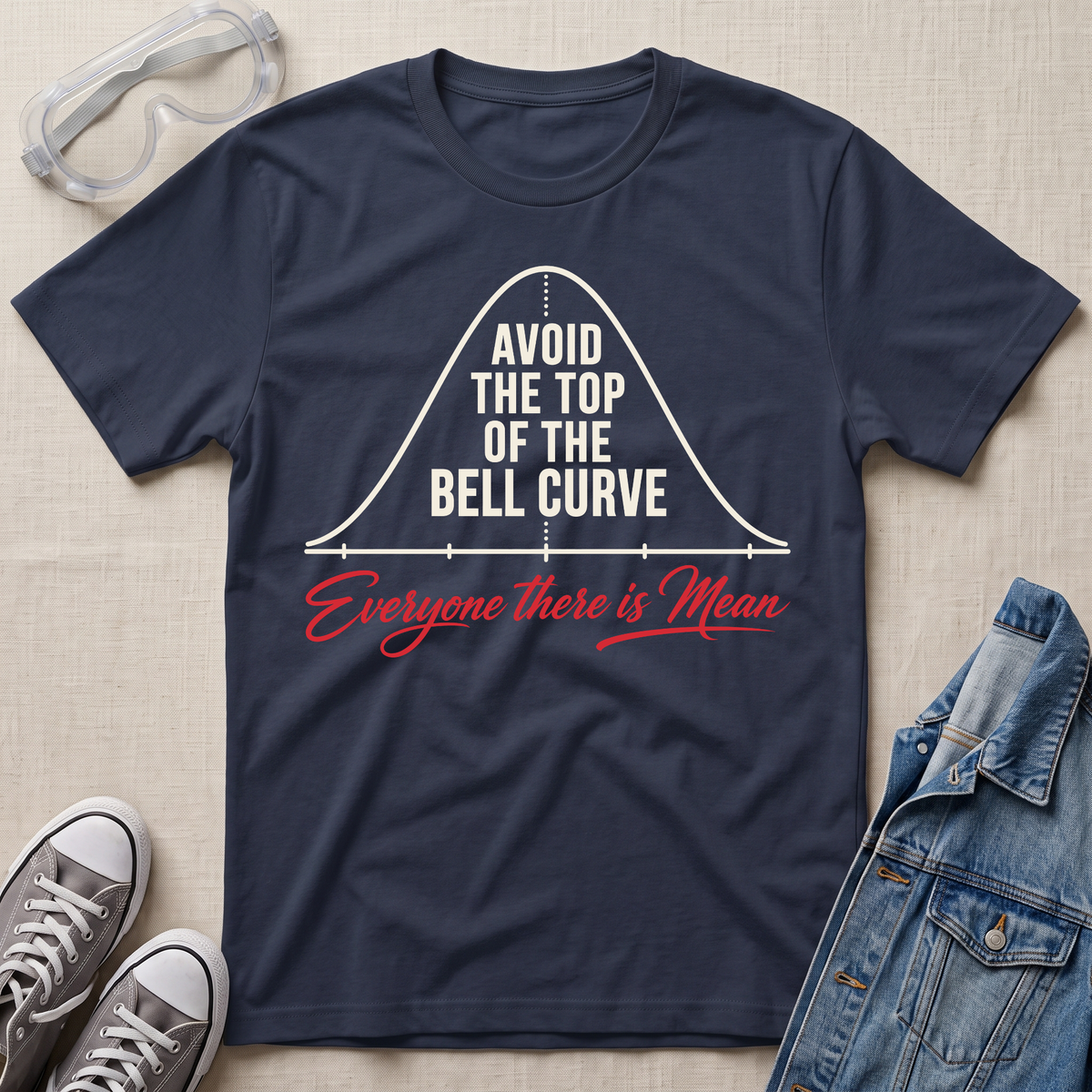 A navy blue graphic t-shirt laid flat on a beige fabric background, styled with safety goggles, gray sneakers, and a denim jacket. The t-shirt features a white bell curve graphic with the text 'AVOID THE TOP OF THE BELL CURVE' and red script text reading 'Everyone there is Mean'.