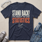 A navy blue graphic t-shirt laid flat on a beige fabric background, surrounded by props including safety goggles, gray sneakers, and a denim jacket. The t-shirt features the text 'STAND BACK! I'M ABOUT TO USE STATISTICS' in white and orange lettering with a small scatterplot graphic below.