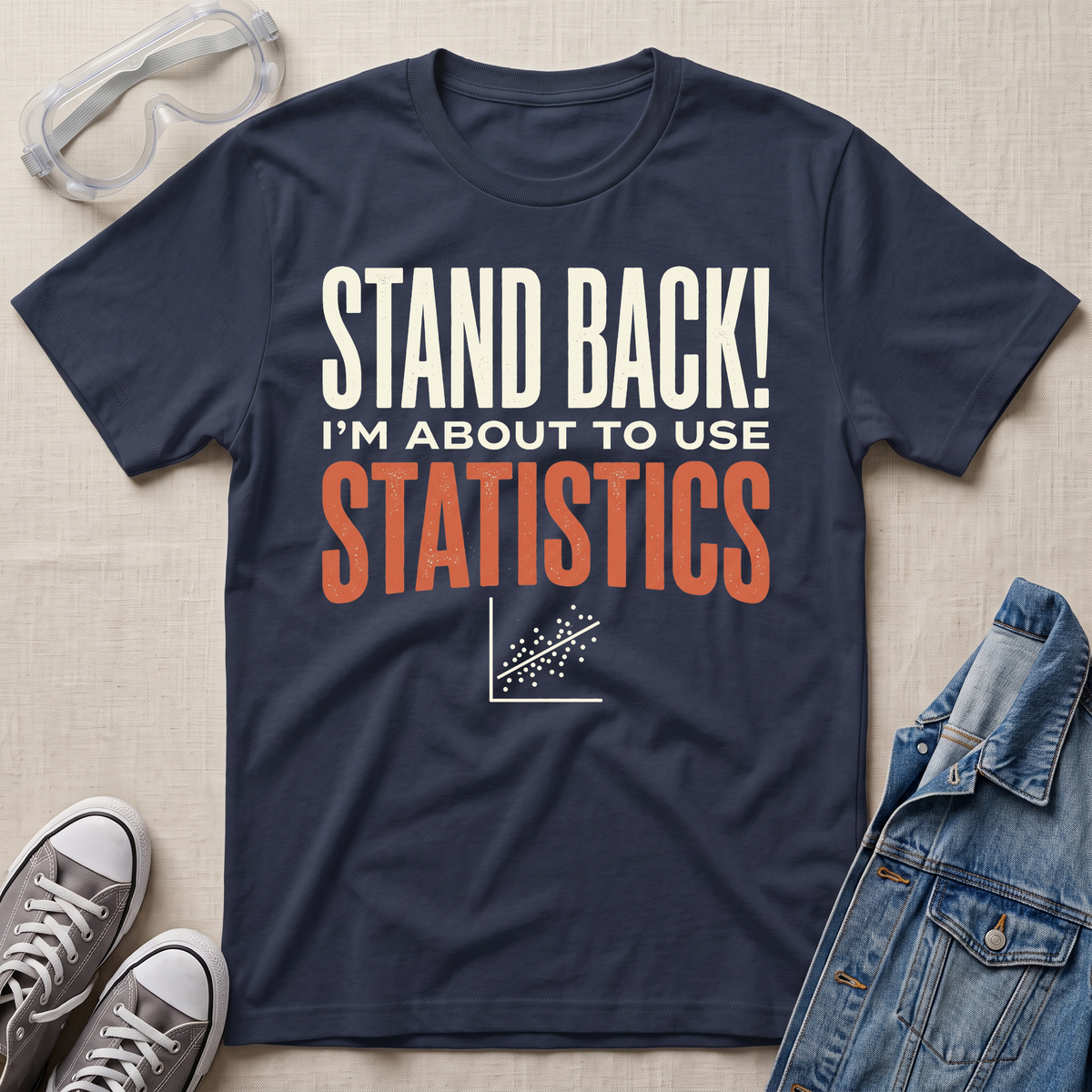 A navy blue graphic t-shirt laid flat on a beige fabric background, surrounded by props including safety goggles, gray sneakers, and a denim jacket. The t-shirt features the text 'STAND BACK! I'M ABOUT TO USE STATISTICS' in white and orange lettering with a small scatterplot graphic below.