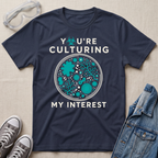 A navy blue graphic t-shirt laid flat on a beige fabric background, surrounded by props including safety goggles, gray sneakers, and a denim jacket. The t-shirt features a circular petri-dish style graphic with teal microorganisms and white text that reads 'YOU'RE CULTURING MY INTEREST'.