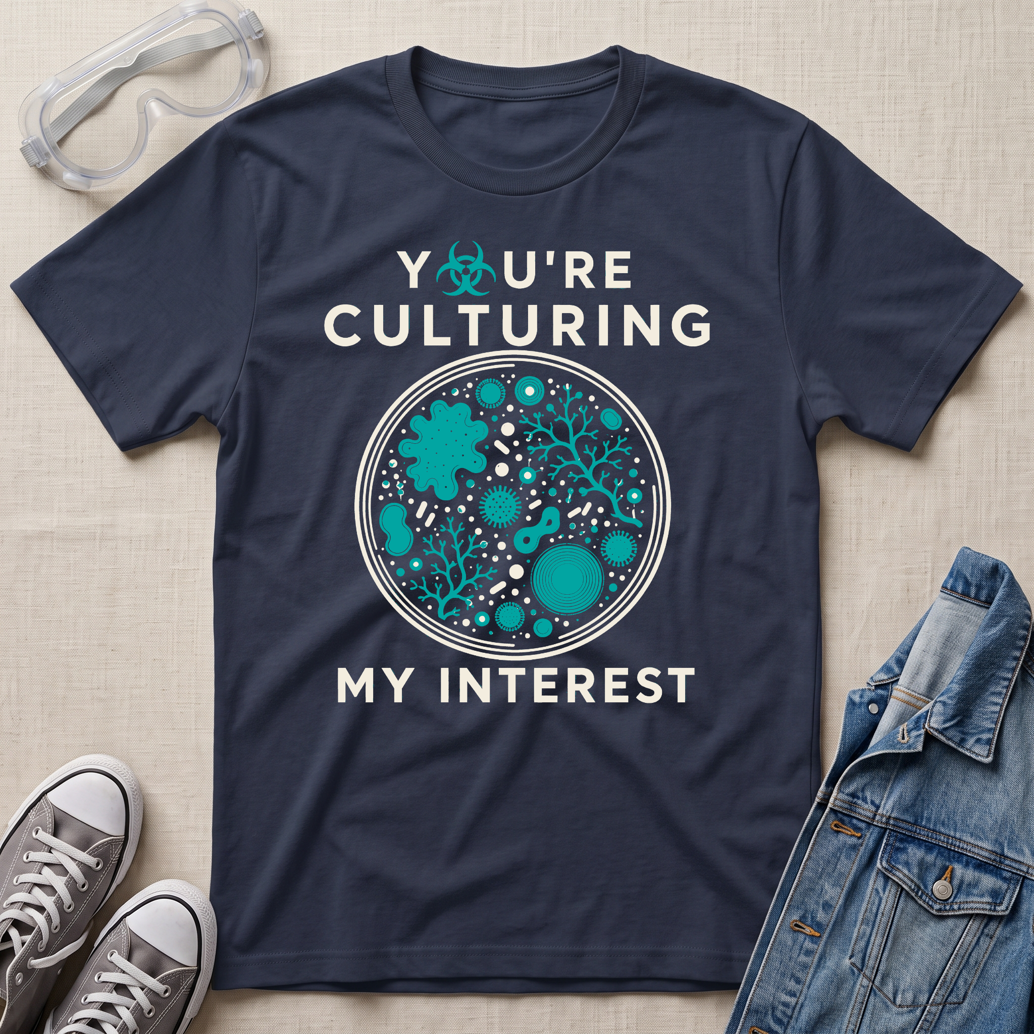 A navy blue graphic t-shirt laid flat on a beige fabric background, surrounded by props including safety goggles, gray sneakers, and a denim jacket. The t-shirt features a circular petri-dish style graphic with teal microorganisms and white text that reads 'YOU'RE CULTURING MY INTEREST'.