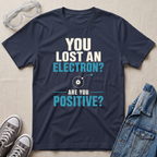 A navy blue graphic t-shirt laid flat on a beige fabric background, surrounded by styling props including safety goggles, gray sneakers, and a denim jacket. The t-shirt features a white and light blue text graphic that reads 'YOU LOST AN ELECTRON? ARE YOU POSITIVE?' with a simple atom diagram.