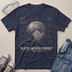 A navy blue graphic T-shirt laid flat on a light beige fabric background, surrounded by styled props including safety goggles, gray sneakers, and a denim jacket. The shirt features a large printed Pluto diagram with the text 'PLUTO: NEVER FORGET 1930-2006' in beige.