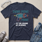 A navy blue graphic t-shirt laid flat on a light beige fabric background, styled with safety goggles, gray sneakers, and a denim jacket. The t-shirt features a colorful retro toy gun illustration with the text 'PEW! PEW! PEW! ... IS THE SOUND OF SCIENCE.'