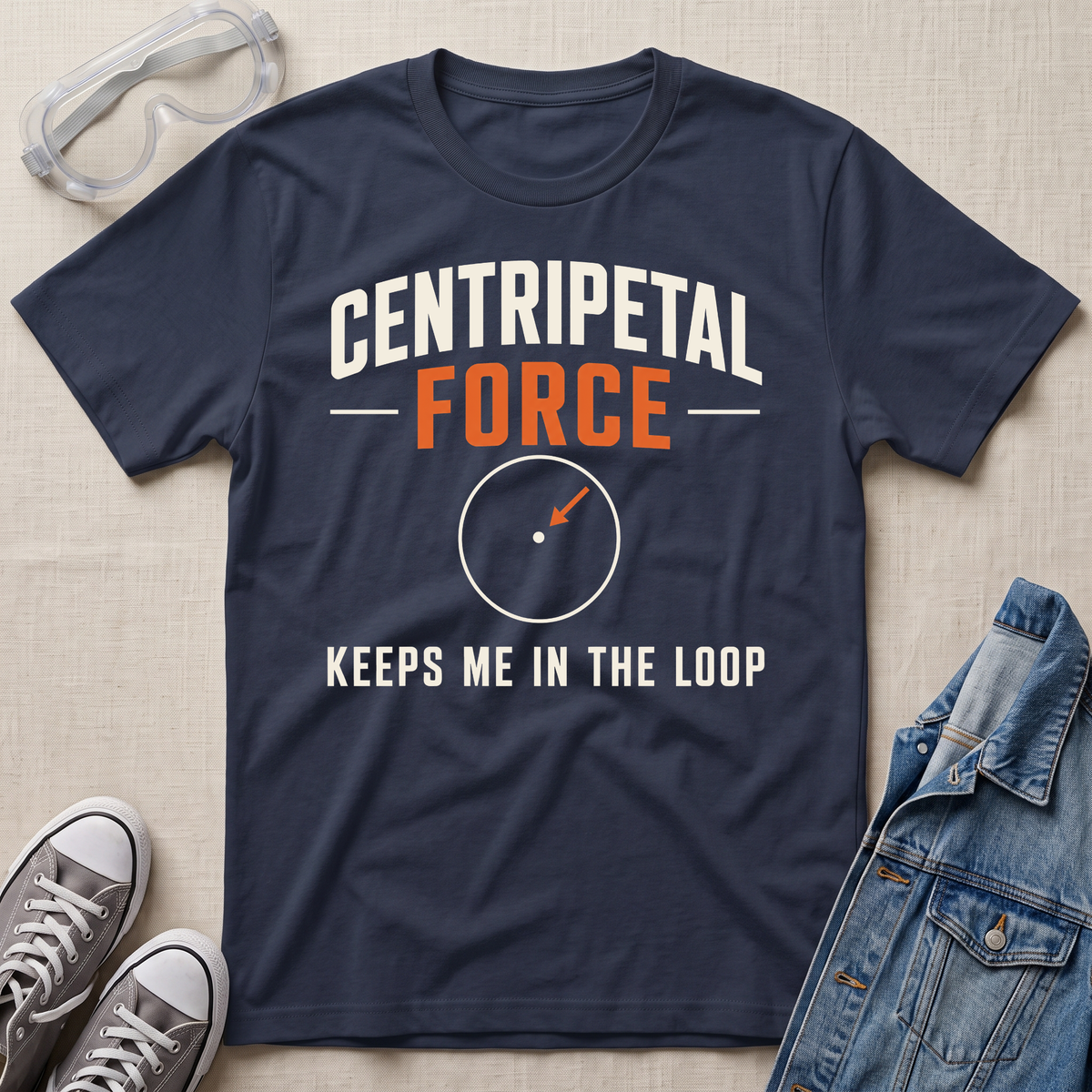 A navy blue graphic t-shirt laid flat on a beige fabric background, styled with safety goggles, gray sneakers, and a denim jacket. The t-shirt features the text 'CENTRIPETAL FORCE' in white and orange, with a circular diagram graphic below it and the phrase 'KEEPS ME IN THE LOOP'.