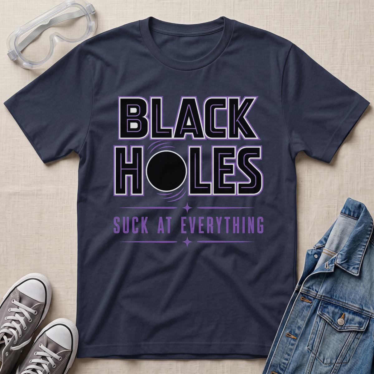 A navy blue graphic t-shirt laid flat on a beige fabric background, styled with safety goggles, gray sneakers, and a denim jacket. The t-shirt features a graphic that reads 'BLACK HOLES SUCK AT EVERYTHING' in black and purple text with a black circle illustration.