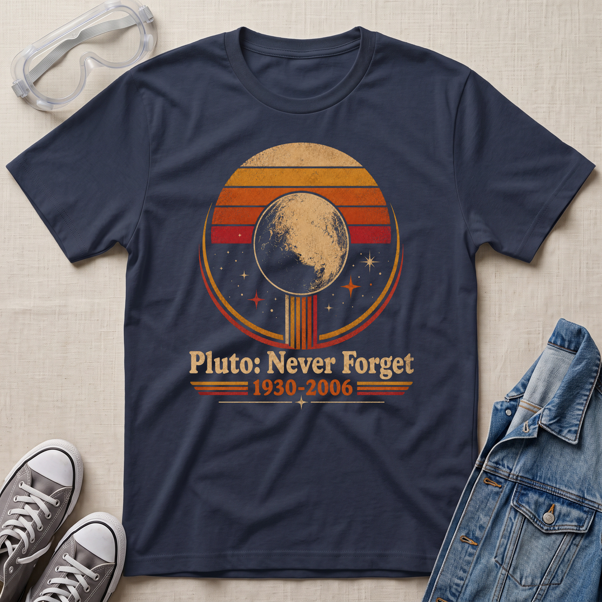 A navy blue graphic t-shirt laid flat on a beige fabric background, styled with clear safety goggles, gray sneakers, and a denim jacket. The t-shirt features a retro circular graphic of Pluto with the text 'Pluto: Never Forget 1930-2006'.