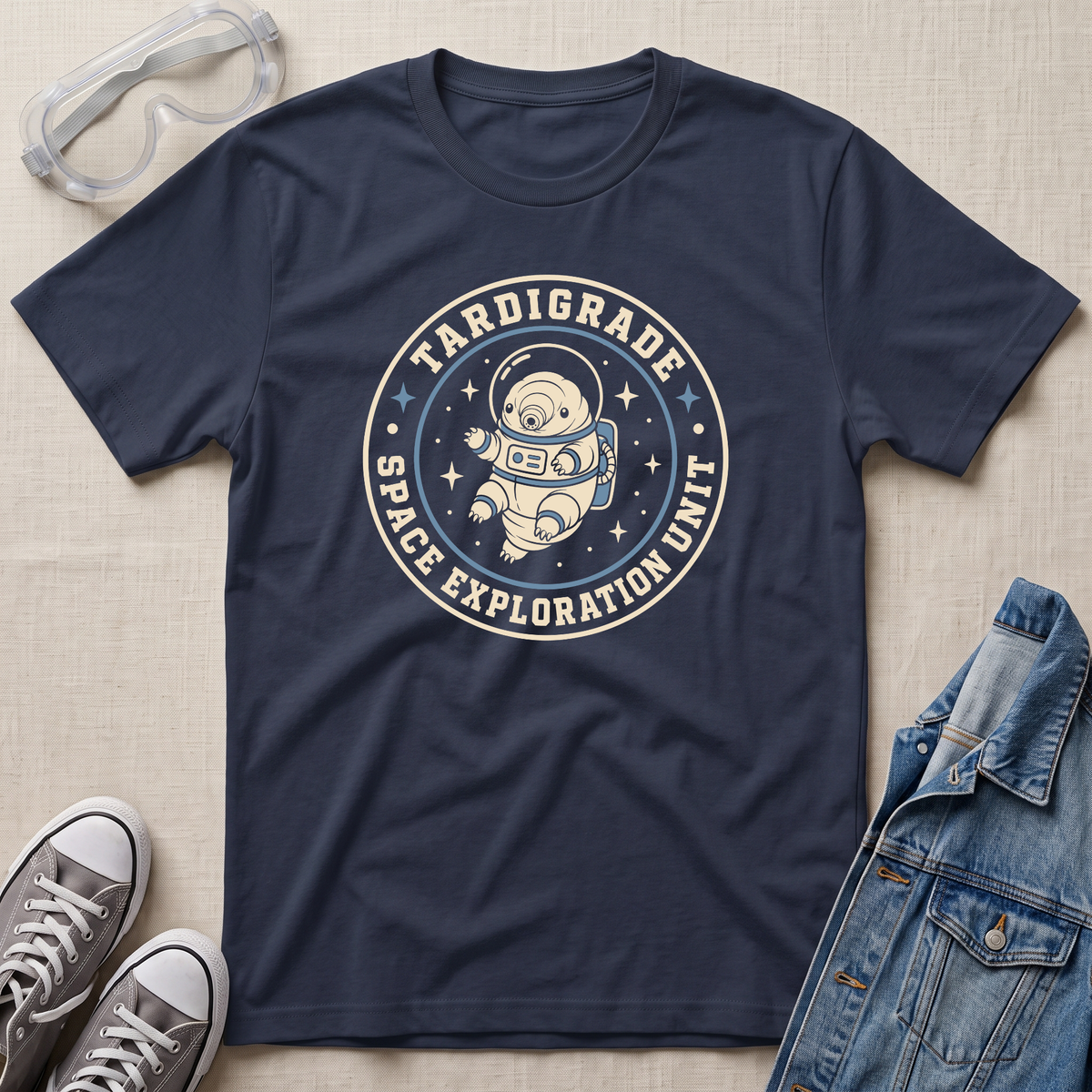 A navy blue graphic T-shirt laid flat on a light beige fabric background, styled with safety goggles, gray sneakers, and a denim jacket. The shirt features a circular retro space-themed graphic reading 'TARDIGRADE SPACE EXPLORATION UNIT' around an astronaut illustration.
