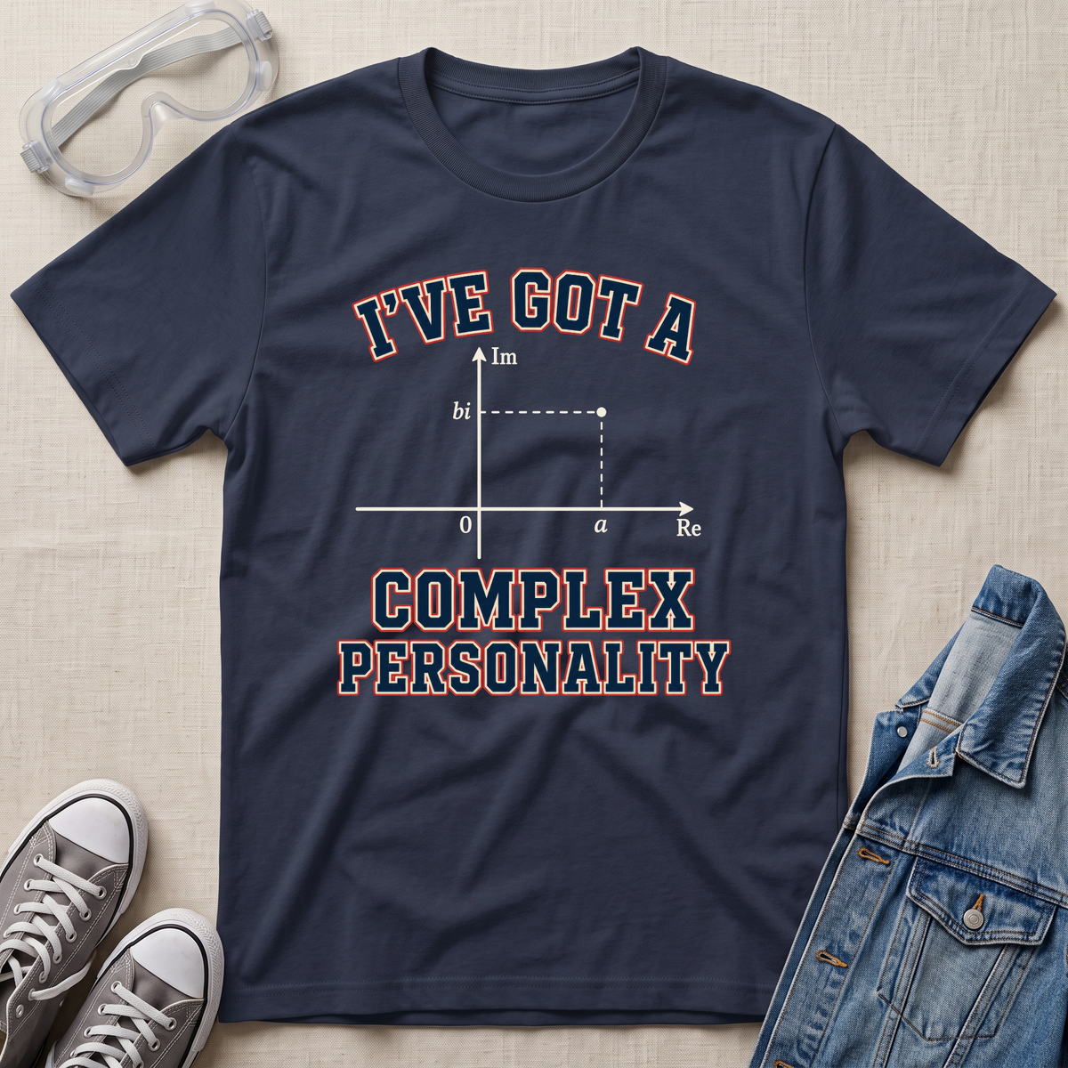 A navy blue graphic t-shirt laid flat on a beige fabric background, styled with safety goggles, gray sneakers, and a denim jacket. The t-shirt features a graphic of a complex number magnitude plot with the text 'I'VE GOT A COMPLEX PERSONALITY' in white with a red outline.