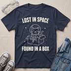 A navy blue  graphic t-shirt laid flat on a beige fabric background, surrounded by styling props including clear safety glasses, gray sneakers, and a blue denim jacket. The t-shirt features a white graphic of a cat inside a box with a helmet and the text 'LOST IN SPACE FOUND IN A BOX'.