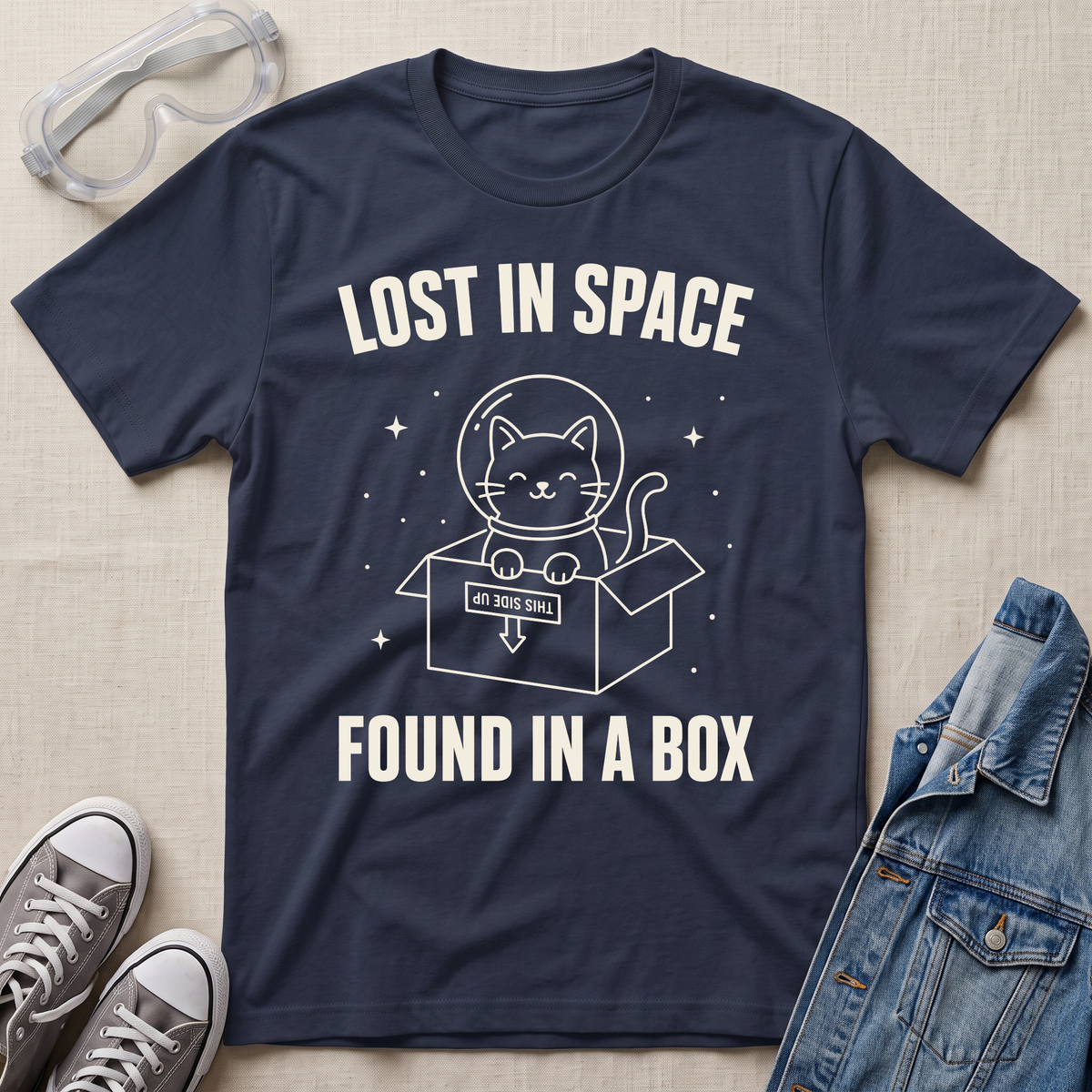 A navy blue  graphic t-shirt laid flat on a beige fabric background, surrounded by styling props including clear safety glasses, gray sneakers, and a blue denim jacket. The t-shirt features a white graphic of a cat inside a box with a helmet and the text 'LOST IN SPACE FOUND IN A BOX'.