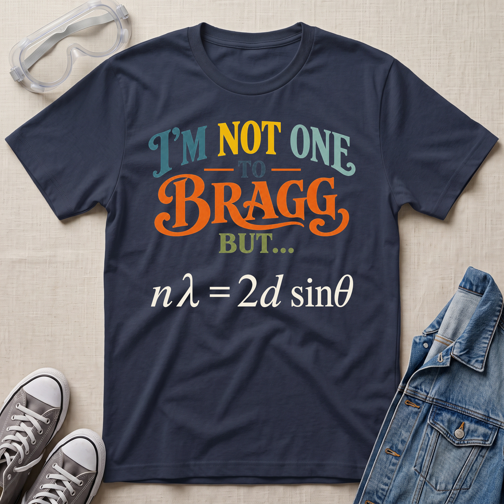 A navy blue graphic t-shirt laid flat on a beige fabric background, styled with safety goggles, gray sneakers, and a denim jacket. The t-shirt features a colorful text graphic reading 'I'M NOT ONE TO BRAGG BUT.