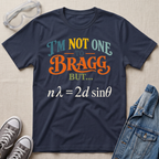A navy blue graphic t-shirt laid flat on a beige fabric background, styled with safety goggles, gray sneakers, and a denim jacket. The t-shirt features a colorful text graphic reading 'I'M NOT ONE TO BRAGG BUT.