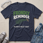 A navy blue graphic t-shirt laid flat on a beige fabric background, surrounded by styling props including safety goggles, gray sneakers, and a denim jacket. The t-shirt features a green fern leaf graphic and the text 'FRONDLY REMINDER PLANTS NEED LIGHT' in green and white.