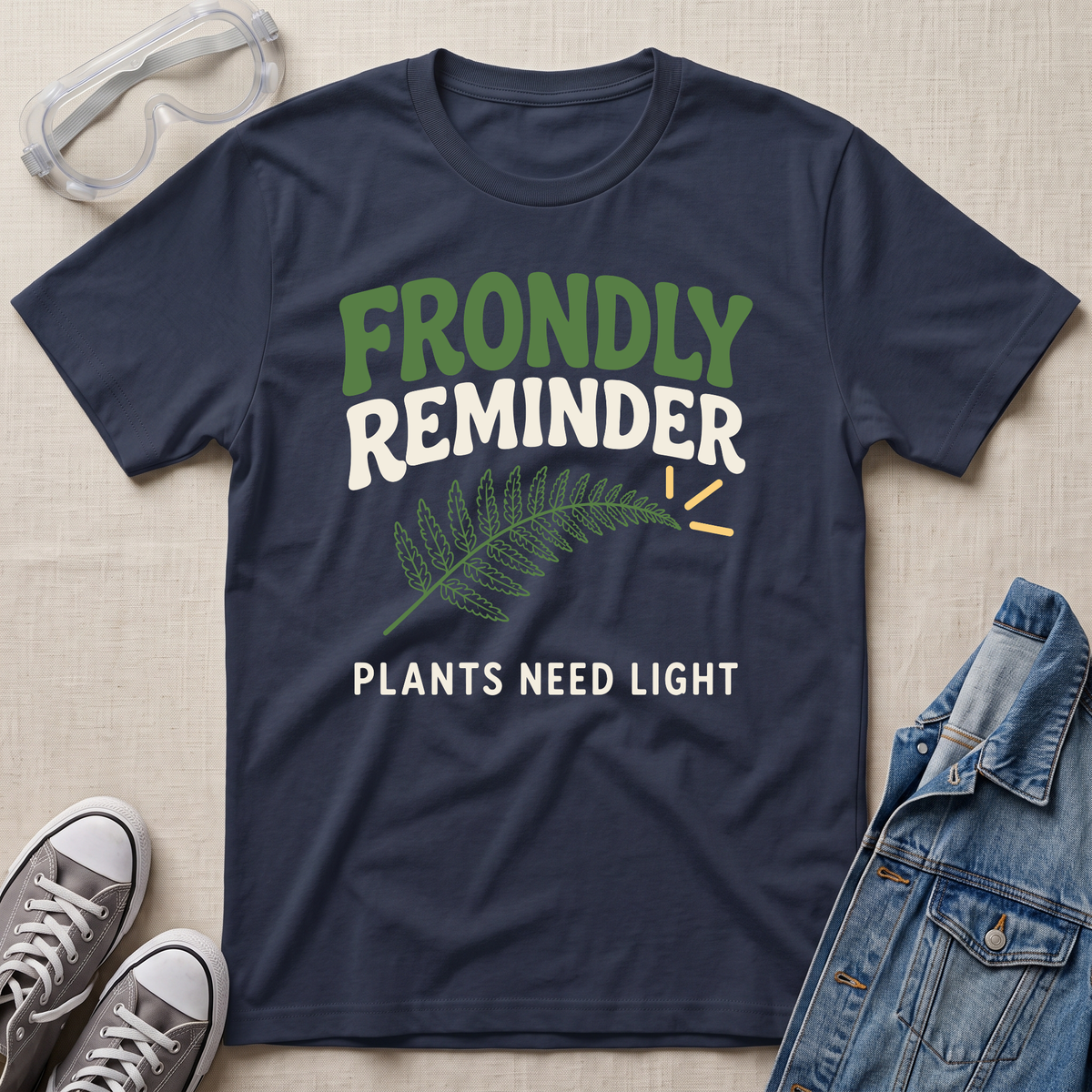 A navy blue graphic t-shirt laid flat on a beige fabric background, surrounded by styling props including safety goggles, gray sneakers, and a denim jacket. The t-shirt features a green fern leaf graphic and the text 'FRONDLY REMINDER PLANTS NEED LIGHT' in green and white.