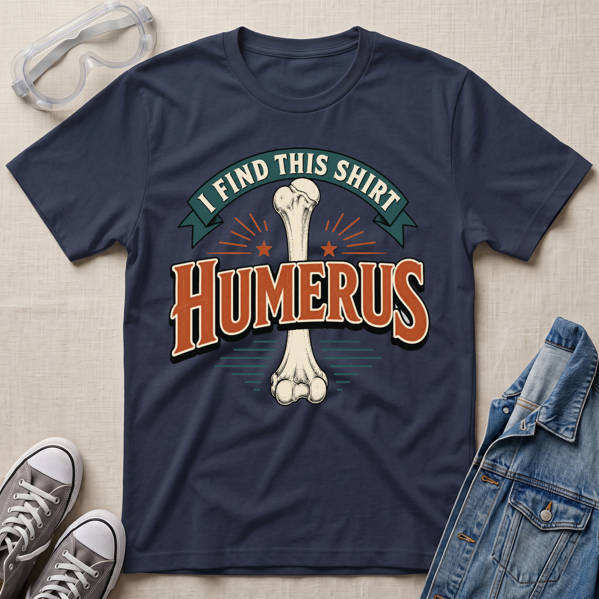 A navy blue graphic t-shirt laid flat on a light wood surface, featuring a bone graphic and the text 'I FIND THIS SHIRT HUMERUS' in orange and green. The shirt is styled with safety glasses, gray sneakers, and a denim jacket.