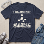 A navy blue graphic t-shirt with white text reading 'I AM A MOLECULE ASK ME ABOUT MY CONFORMATION' and a simple molecule model printed on the front. The shirt is displayed in a flat-lay arrangement on a beige fabric background, styled with safety goggles, gray sneakers, and a denim jacket.