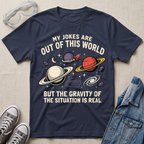 A navy blue graphic t-shirt laid flat on a light beige fabric background, surrounded by styling props including clear safety glasses, gray sneakers, and a blue denim jacket. The t-shirt features a space-themed graphic with planets, stars, and the text 'MY JOKES ARE OUT OF THIS WORLD BUT THE GRAVITY OF THE SITUATION IS REAL'.