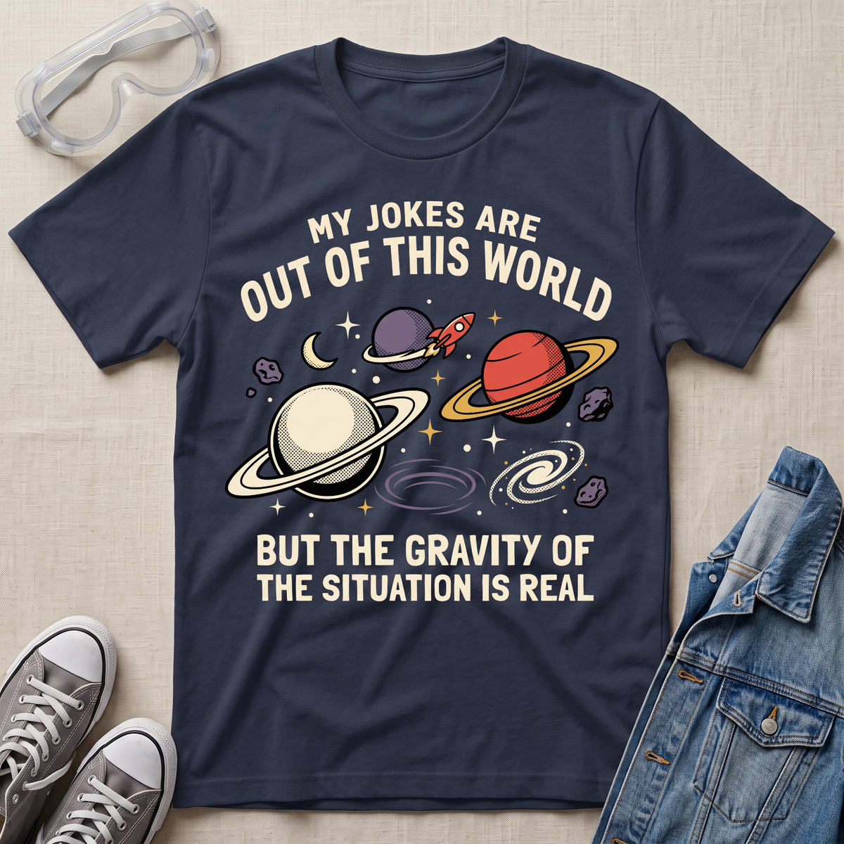A navy blue graphic t-shirt laid flat on a light beige fabric background, surrounded by styling props including clear safety glasses, gray sneakers, and a blue denim jacket. The t-shirt features a space-themed graphic with planets, stars, and the text 'MY JOKES ARE OUT OF THIS WORLD BUT THE GRAVITY OF THE SITUATION IS REAL'.
