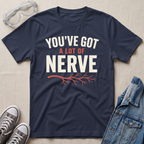 A navy blue graphic t-shirt laid flat on a beige fabric background, styled with safety goggles, gray sneakers, and a denim jacket. The t-shirt features the text 'YOU'VE GOT A LOT OF NERVE' in white and orange lettering with an orange branch graphic.