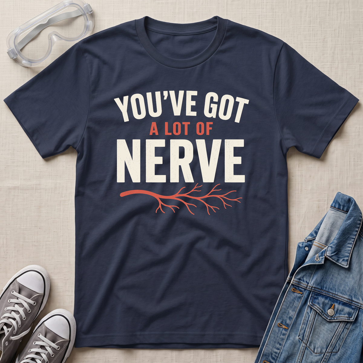 A navy blue graphic t-shirt laid flat on a beige fabric background, styled with safety goggles, gray sneakers, and a denim jacket. The t-shirt features the text 'YOU'VE GOT A LOT OF NERVE' in white and orange lettering with an orange branch graphic.