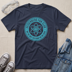 A navy blue graphic t-shirt laid flat on a beige fabric background, styled with safety goggles, gray sneakers, and a denim jacket. The t-shirt features a teal circular Heisenberg fan club graphic with an atom symbol and the text 'HEISENBERG FAN CLUB' and 'WE'RE NOT CERTAIN WE'RE HERE.