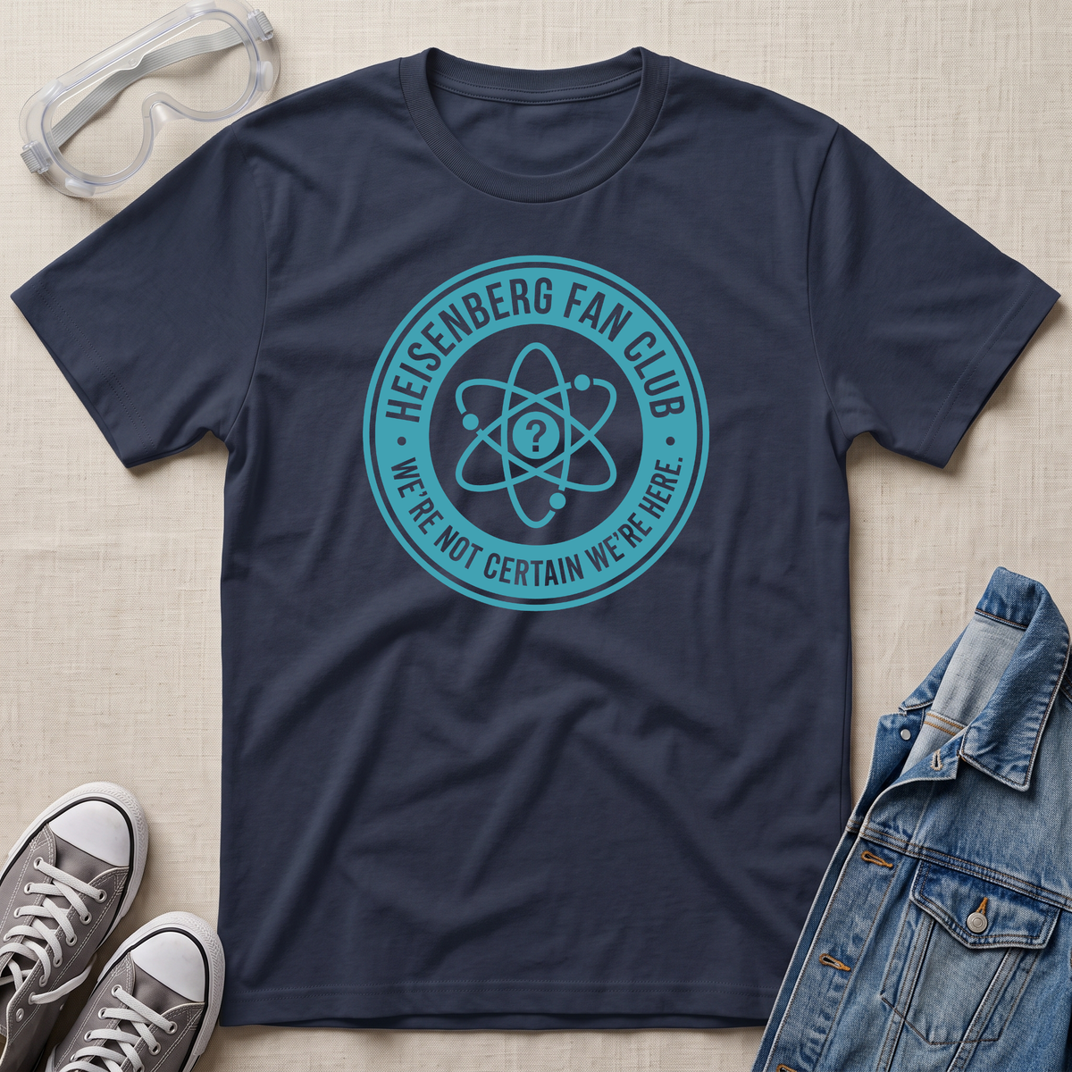 A navy blue graphic t-shirt laid flat on a beige fabric background, styled with safety goggles, gray sneakers, and a denim jacket. The t-shirt features a teal circular Heisenberg fan club graphic with an atom symbol and the text 'HEISENBERG FAN CLUB' and 'WE'RE NOT CERTAIN WE'RE HERE.