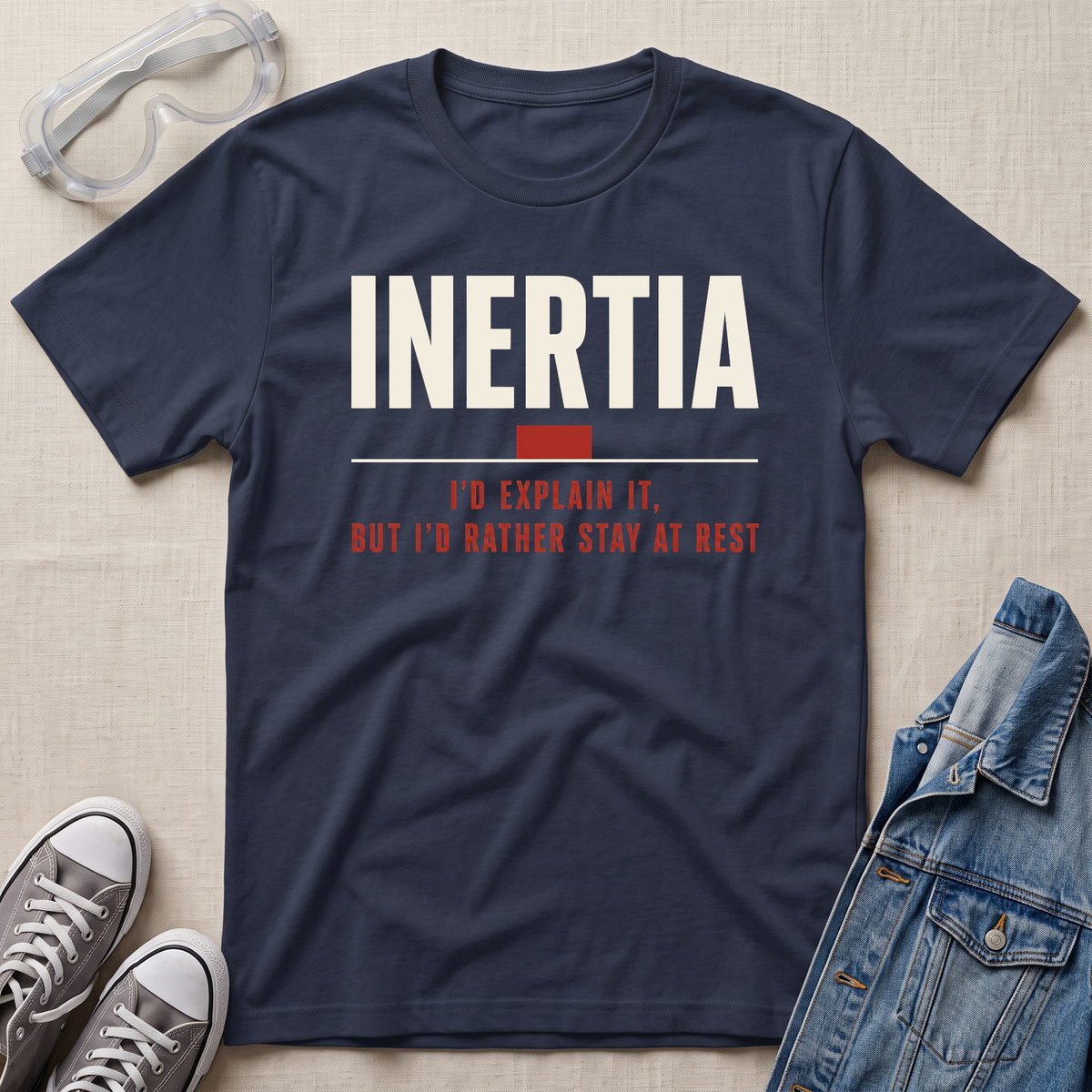 A navy blue graphic t-shirt laid flat on a beige fabric background, styled with safety goggles, gray sneakers, and a denim jacket. The t-shirt features the word 'INERTIA' in bold white letters with a red block and red text underneath reading 'I'D EXPLAIN IT, BUT I'D RATHER STAY AT REST'.