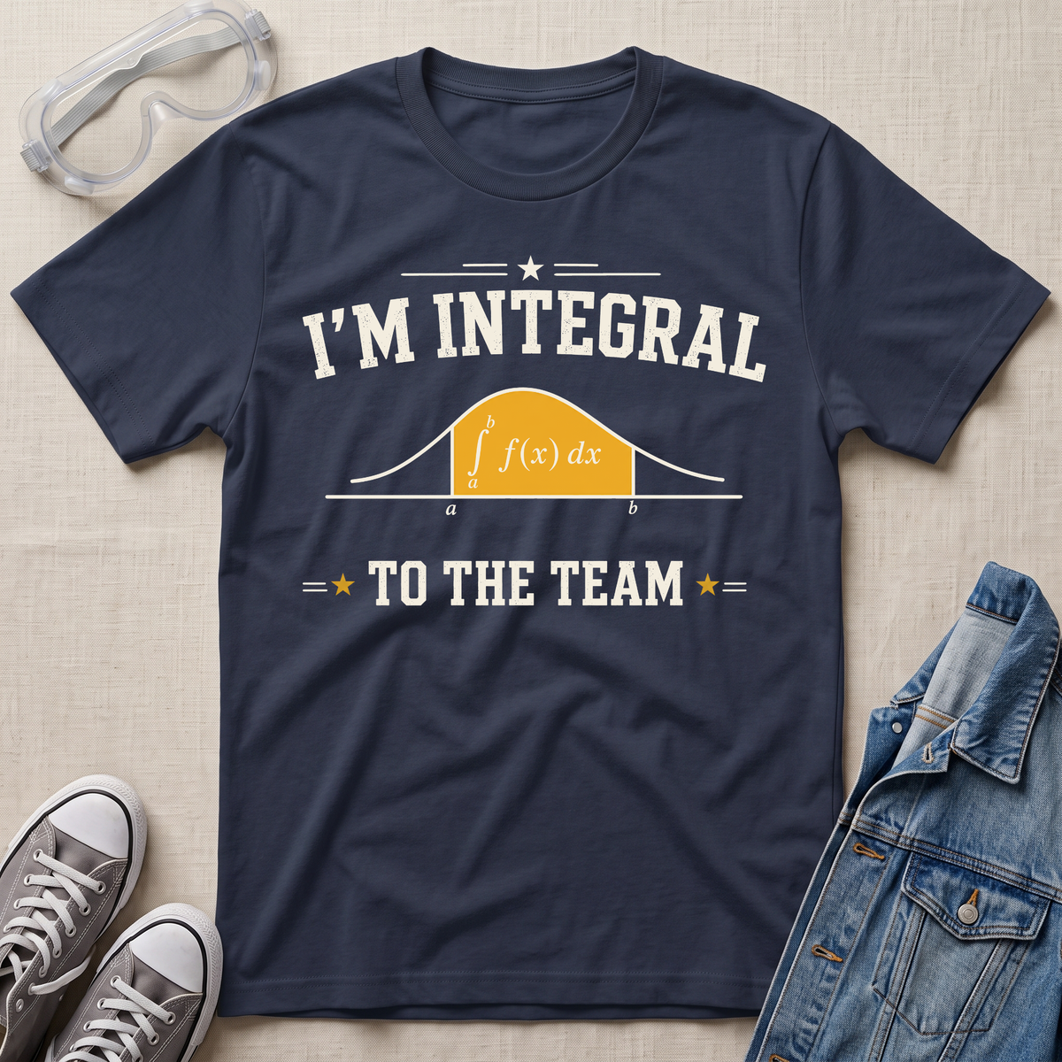 A navy blue graphic t-shirt laid flat on a beige fabric background, styled with safety goggles, gray sneakers, and a denim jacket. The t-shirt features a white graphic that reads 'I'M INTEGRAL TO THE TEAM' with a yellow integral curve diagram.