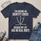 A navy blue graphic t-shirt laid flat on a light beige fabric background, surrounded by styling props including safety goggles, gray sneakers, and a denim jacket. The t-shirt features a white printed graphic of a parabola and the text 'I'M HAVING AN IDENTITY CRISIS BECAUSE MY LIFE HAS NO REAL ROOTS'.