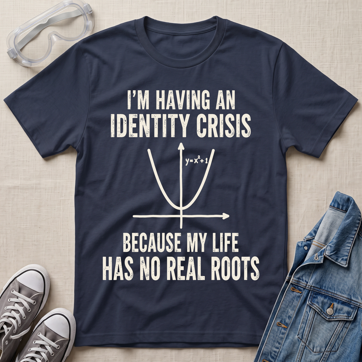 A navy blue graphic t-shirt laid flat on a light beige fabric background, surrounded by styling props including safety goggles, gray sneakers, and a denim jacket. The t-shirt features a white printed graphic of a parabola and the text 'I'M HAVING AN IDENTITY CRISIS BECAUSE MY LIFE HAS NO REAL ROOTS'.