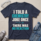 A navy blue graphic t-shirt with the text 'I TOLD A CHEMISTRY JOKE ONCE THERE WAS NO REACTION' in white and teal lettering, styled in a flat-lay with safety goggles, sneakers, and a denim jacket on a beige fabric background.