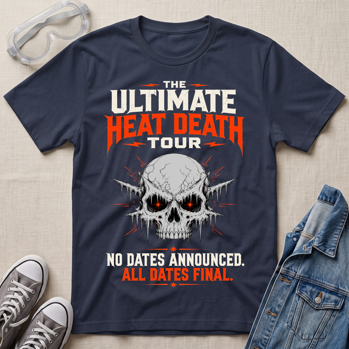 A navy blue graphic t-shirt laid flat on a beige fabric background, styled with safety goggles, gray sneakers, and a denim jacket. The t-shirt features a skull graphic with glowing red eyes and bold text that reads 'THE ULTIMATE HEAT DEATH TOUR' and 'NO DATES ANNOUNCED.