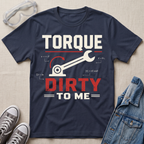 A navy blue graphic t-shirt laid flat on a beige fabric background, styled with safety glasses, gray sneakers, and a denim jacket. The t-shirt features a white and red graphic that reads 'TORQUE DIRTY TO ME' with a wrench and torque equation illustration.
