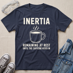 A navy blue graphic t-shirt laid flat on a beige fabric background, surrounded by styling props including safety goggles, gray sneakers, and a denim jacket. The t-shirt features a white graphic of a steaming coffee mug with the text 'INERTIA REMAINING AT REST UNTIL THE CAFFEINE KICKS IN'.