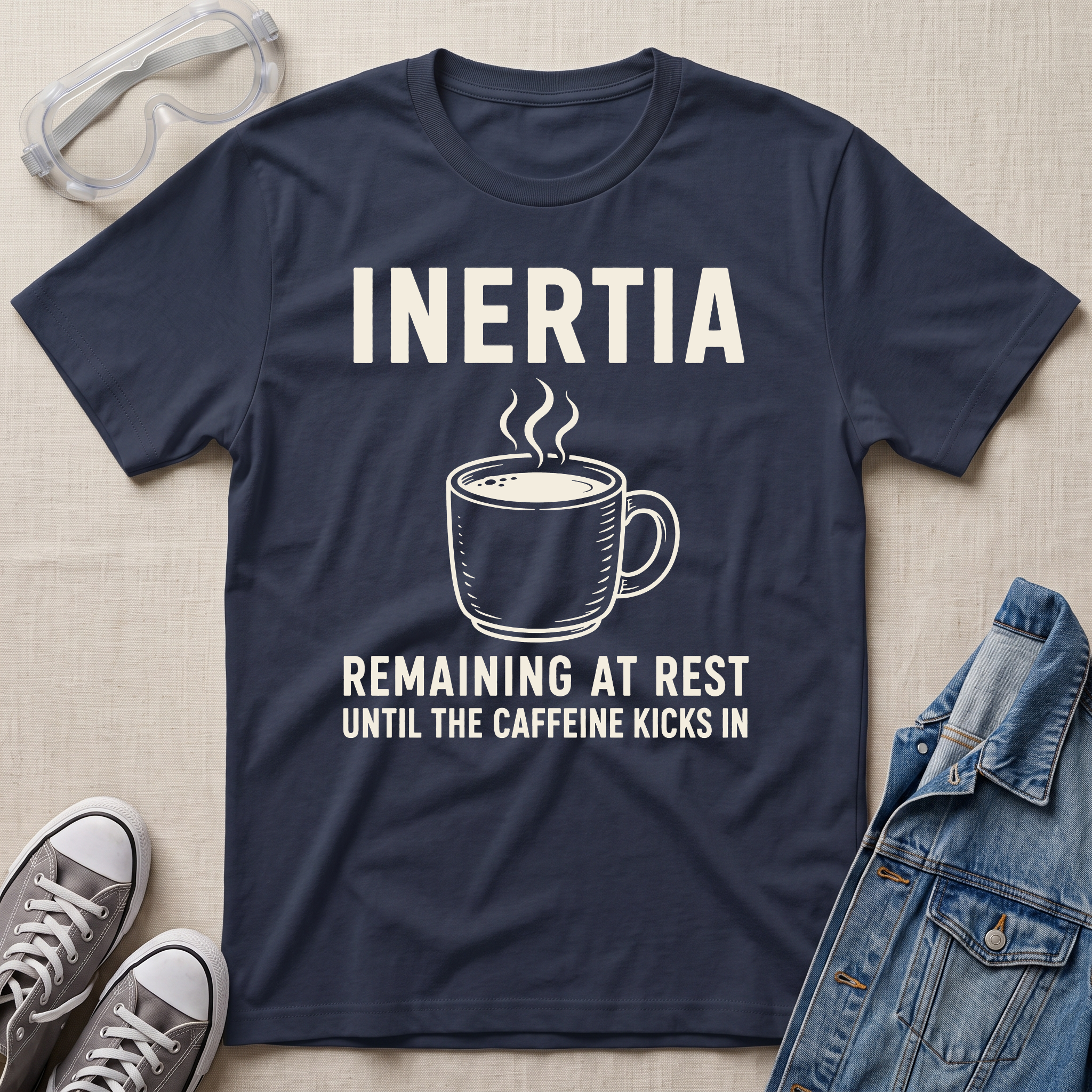A navy blue graphic t-shirt laid flat on a beige fabric background, surrounded by styling props including safety goggles, gray sneakers, and a denim jacket. The t-shirt features a white graphic of a steaming coffee mug with the text 'INERTIA REMAINING AT REST UNTIL THE CAFFEINE KICKS IN'.