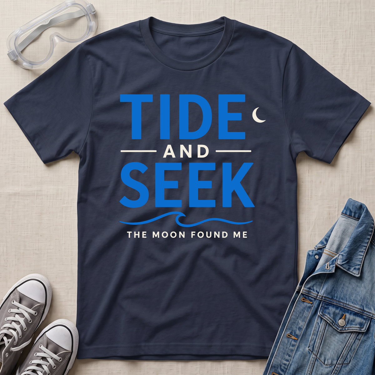 A navy short-sleeve t-shirt with a blue graphic reading 'TIDE AND SEEK THE MOON FOUND ME' laid flat on a beige textured surface, surrounded by styling props including safety goggles, gray sneakers, and a denim jacket.
