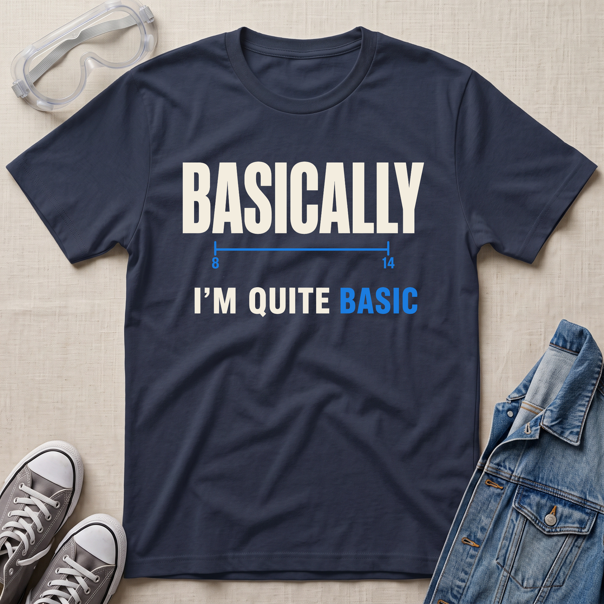 A navy blue graphic t-shirt laid flat on a beige fabric background, styled with safety goggles, gray sneakers, and a denim jacket. The t-shirt features the text 'BASICALLY I'M QUITE BASIC' in white and blue lettering.