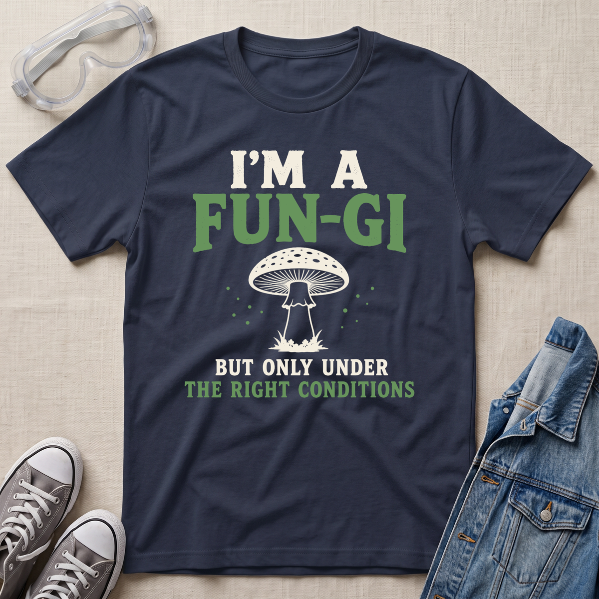 A navy blue graphic t-shirt laid flat on a beige fabric background, styled with safety goggles, gray sneakers, and a denim jacket. The t-shirt features a white mushroom illustration and the text 'I'M A FUN-GI BUT ONLY UNDER THE RIGHT CONDITIONS' in white and green.