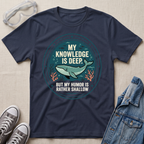 A navy blue graphic t-shirt laid flat on a beige fabric background, styled with safety goggles, gray sneakers, and a denim jacket. The t-shirt features a circular graphic with a cartoon whale and the text  'MY KNOWLEDGE IS DEEP BUT MY HUMOR IS RATHER SHALLOW'.