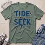 A sage green short-sleeve graphic T-shirt laid flat on a light beige textured surface, with safety glasses, gray sneakers, and a folded denim jacket around it. The shirt has large navy text reading 'TIDE AND SEEK' with smaller text 'THE MOON FOUND ME' and a crescent moon graphic.